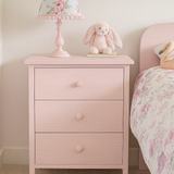 Pink nightstand with floral lamp and stuffed animal, next to a bed with floral bedding, in a child's bedroom.
