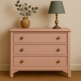 Peachy Keen dresser with three drawers, a vase with greenery, and a lamp on top against a beige wall.