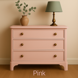 Pink dresser with gold knobs against a beige wall, featuring a lamp with a sage lamp shade and plant decor.